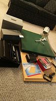 Photo showing guillotine paper cutter, metal storage boxes, file tray, alarm clock, desk lamp, bulletin board, and various small office supplies spread out on carpet.