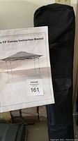 Black storage bag standing upright with a printed instruction manual in front showing the 13'x13' canopy picture and text.