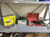 Full shelf view showing boxed corner clamp, Motomaster battery charger, red plastic soldering accessory case, soldering station with coil stand, and corner clamps.