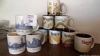 Photo showing ten Starbucks city and destination themed collector mugs stacked and lined up on a surface, with visible labels and designs from multiple locations.