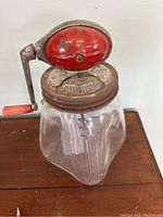 Front view of the hand crank butter churn showing the clear glass jar, metal lid, red crank handle, and the internal metal paddles for churning.