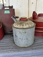 Silver-tone corrugated cylindrical metal gas/oil can with spout and handle, and two red metal cans (round and tall rectangular) in background