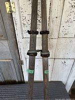 Photo showing two antique rustic wooden oars leaning against a distressed white peeling door. Each oar has metal clamps and bands around the midsection.