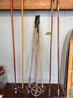 Four antique wooden ski poles standing vertically against a wall, showing natural wood finish and metal baskets at bottom, used for winter sports decor.