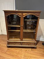 Front view of antique walnut china cabinet showing glass double doors, brass round knobs, and carved ornamental detail at the top and bottom front edges. The cabinet stands on short feet.