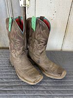 Pair of brown leather Ariat cowgirl boots standing upright showing full side and front view with visible colorful stitching on the shaft and square toe shape.
