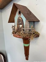 Side view showing birdhouse with distressed blue body, peaked roof and natural moss with miniature wooden fence and bird ornament. Mounted on polished wooden stand fixed to wall.
