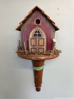 Front view of handcrafted decorative birdhouse mounted on turned wooden base, showing arched door, circular entry hole, and moss with twig fencing detail.