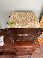 Closed view of foot stool showing patterned upholstered cushioned top and carved wooden body with decorative accents.