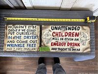 Two rectangular hand-painted wooden signs with humorous sayings on a gray cloth surface with a measuring tape showing approximately 16 inches in length.