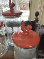 Close-up of 2 red rooster lid canisters, showing the rooster handle detail and the glass body.