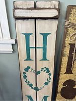 Close front view of a hand painted wooden sign with cream background, teal letters 'H','M','E' and a heart wreath in place of 'O'.