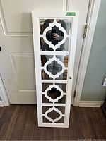 Photo of a white wall hung jewellery cabinet with a mirrored front door featuring a decorative cut-out design, shown closed and standing upright against a door.
