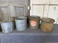 Four minnow buckets standing side by side, two brighter silver color and two rusted, displayed on a cloth surface against a rustic background.
