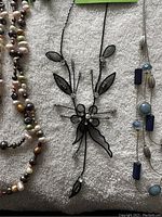 Close view of a black metal wire necklace with leaf and flower design and a silver-tone necklace with blue and round stone beads with crystal accents beside multi-colored freshwater pearl strands.