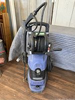 Front view of blue and black Simoni power washer with hose reel and spray gun mounted on wooden floor.