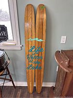 Full view of the pair of vintage wooden water skis standing upright against a wall, showing blue vinyl lettering and wave graphic.