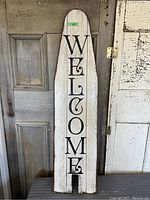 Front view of the antique wooden ironing board painted with 'WELCOME' vertically in black on a white distressed surface.