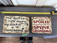 Photo showing two wooden signs side by side with measuring tape above them showing size, and floor visible below with person's feet.