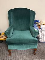 Front view of green velveteen upholstered wingback armchair with wooden legs on carpeted floor.