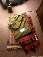 Three assorted throw blankets stacked on carpet, showing solid green throw on top, red and black plaid in the middle, and multi-color patterned throw on the bottom.
