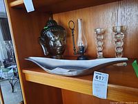 Shelf with glass Buddha head statue, glass oil lamp with black metal base and handle, metal decorative dish, and two glass candle sticks.
