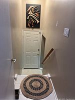 Photo showing carved wood plaque mounted high on a wall above a door and a round woven straw rug on the floor at the base of stairs.