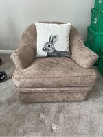 Front view of tan upholstered armchair with decorative rabbit cushion on a carpeted floor.