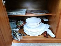 Shelving unit with stacked white porcelain bowls and plates, forks with brown handles, napkin rings wrapped in paper, clear glass plate, and box labeled 'melamine table mats'