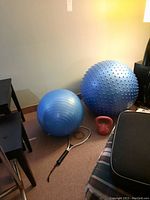 Large blue textured pilates ball, medium blue smooth pilates ball, red kettlebell weight, and a stringed racket with black grip sitting on carpet next to furniture.