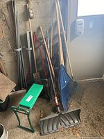 Overall view of the lot showing a variety of gardening tools leaning against the wall including rakes, shovel, hoe, edger, and the green foldable bench/kneeling pad