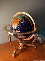 Front angle view of the decorative world globe showing colorful stone inlaid continents, dark blue enamel ocean, brass equator and supporting stand.
