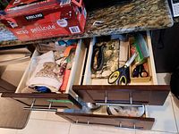 Top view of two kitchen drawers; left drawer contains assorted kitchen towels and napkins, right drawer includes scissors, a vegetable peeler, and resealable bags.