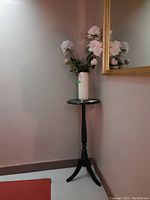 Full view of black wood round table stand with ceramic vase holding faux pink roses against wall and mirror