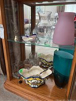 Showcase of glass vases, ceramic bowl, brass bell, trinket dish, paperweight, rose quartz and glass egg on glass shelf inside wood cabinet.