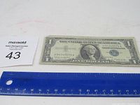 1957 B $1 Silver Certificate Blue Seal bill laid flat showing full front side alongside a blue ruler for scale and a MaxSold auction card with lot number.
