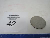 1971 Eisenhower silver-colored dollar coin placed on a white surface with a maxsold auction lot number 42 card nearby, showing obverse side with Dwight D. Eisenhower profile portrait.