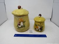 Front view of large and small yellow ceramic canisters with mushroom decorations and lids with mushroom knobs.