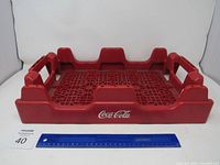 Red Coca-Cola bottle case front view with ruler for scale, showing grid base and side handles.