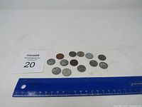 Top view of 12 steel penny coins laid out on a white background with a blue ruler beneath them for scale. The coins vary in wear and color.