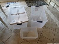 Three clear plastic containers on tiled floor: one small container without lid, two larger containers with white lids and black handles.
