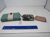 Three purses including teal and white wallet, black and yellow floral wristlet, and pink wallet laid side by side.