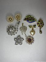 Photo showing a variety of vintage assorted brooches, pins, and earrings with rhinestones, faux pearls, and colorful stones arranged on a gray surface.