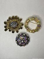 Lot of three assorted vintage brooches including flower-shaped gold-tone with blue crystals, floral wreath with pastel rhinestones, and round black-tone floral brooch with multi-color crystals.