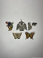Six assorted jewelry pins and brooches on white background including rooster, thunderbird, otter, bear, and two butterflies.