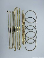 Photo showing six gold tone bangles arranged vertically with five gold tone chain style bracelets with flat ID plates beside them on white background.
