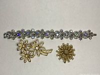 Silver-tone bracelet with linked floral and bead designs accented with purple and clear rhinestones, displayed next to two gold-tone brooches with pearl-like bead details.
