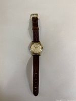 Full length view of brown leather strap wristwatch with gold-tone case and white dial.