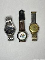 Three watches laid flat showing faces and bands: silver-tone with dark dial, Boyz n the Hood branded watch with leather band, and gold-tone watch with metal band.
