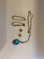 Sterling silver necklace with blue-green mother of pearl pendant and two pairs of earrings: knot studs and chain tassel earrings, arranged on a surface.